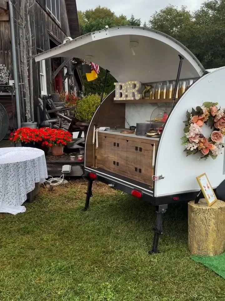 The trailer open for service on the lawn of a barn wedding, with a wreath, red flowers and a lace-covered table