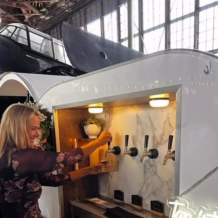 A host pours a drink from one of the four taps inside the lit trailer at an indoor event