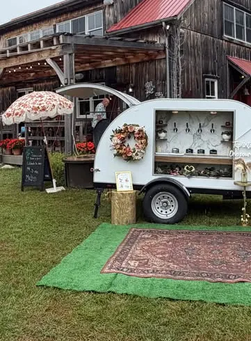 The tap trailer on a lawn in front of a rustic barn, with a vintage rug, a floral umbrella and a wreath on the trailer