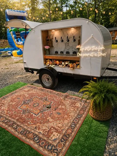 The Sticks & Stems tap trailer set up outdoors on a vintage rug, with string lights, a fern and the neon sign reading tap into the fun